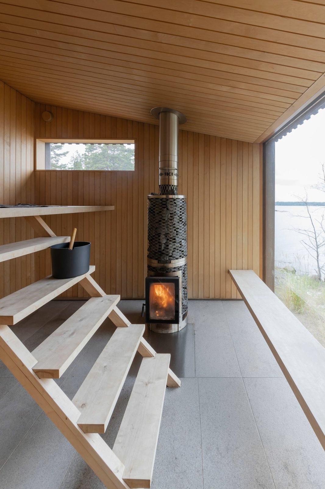 A timber sauna with a lit wood stove and a long window onto the sea
